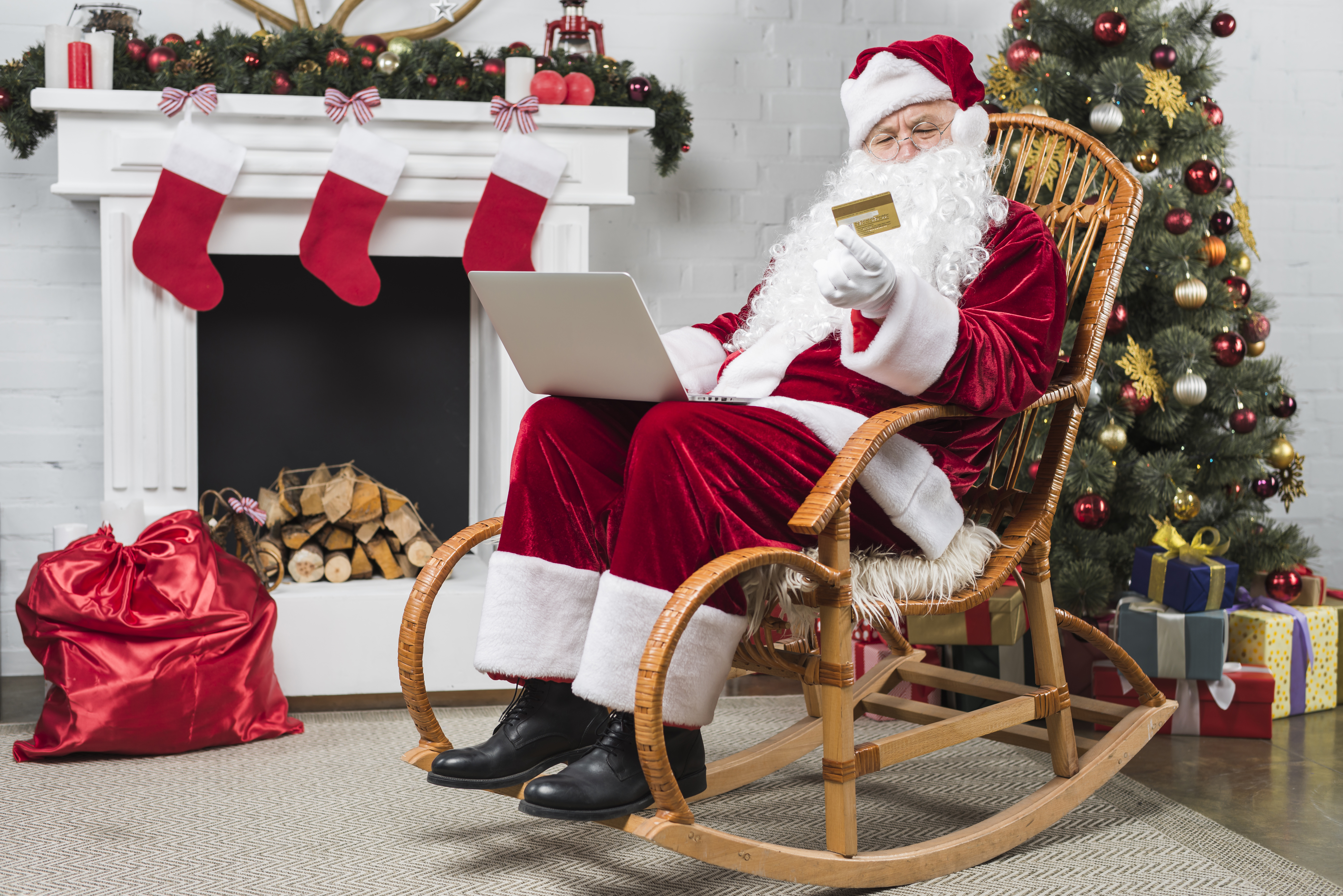 Santa sitting on a rocking chair next to a Christmas tree and fireplace online Christmas shopping on a laptop with a credit card